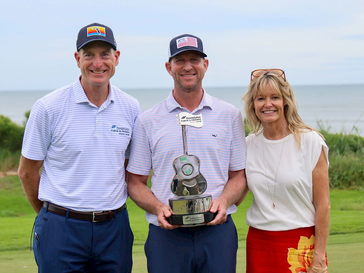 Three golfers in caps and striped polos pose outdoors by the ocean, holding a large award trophy.