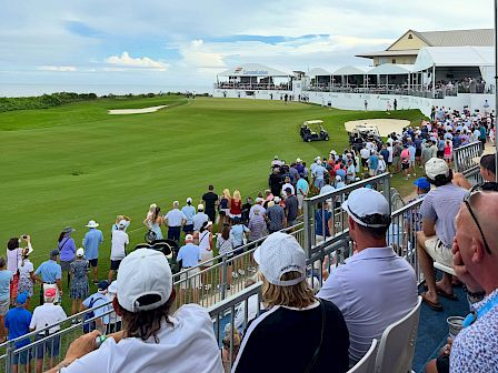A packed golf tournament on a seaside course, with players on the green, spectators in stands, and sponsor tents in view.