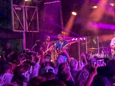 A live rock concert&mdash;guitarists onstage under purple spotlights and fog, with the crowd cheering in front of glowing LED lights.