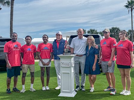 A group in red shirts stands on a grassy field at an outdoor event, posing with a trophy in front of a white van.