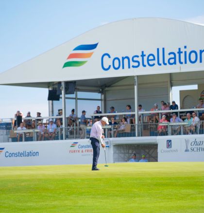 A golfer tees off on a green at a tournament, with a large &ldquo;Constellation&rdquo; tent and spectators in the background.