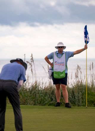A golfer in a cap and vest waves a flag on a golf course while a photographer watches under cloudy skies.