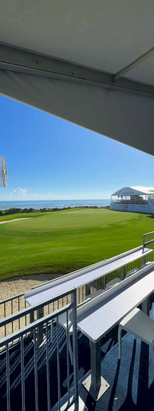 A sunny seaside golf course viewed from a covered terrace, with patio chairs, a scoreboard screen, and the ocean horizon.
