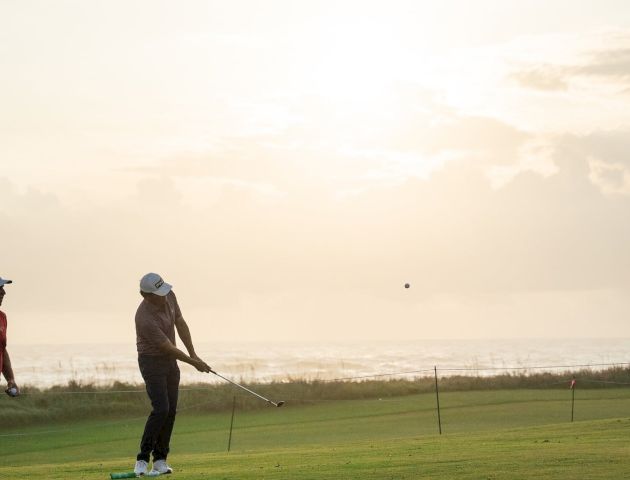 A golfer in a cap swings on a green course toward a distant ball under a cloudy sky.
