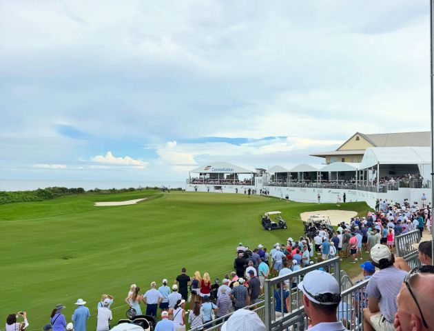 A packed golf tournament at a seaside course, with players on the green and tents by the clubhouse under a cloudy sky.