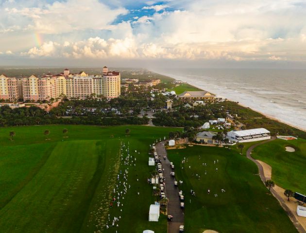 Aerial view of a coastal resort with manicured golf course, beachfront buildings, and dramatic clouds over the ocean.