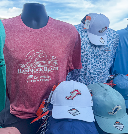 A display of assorted hats and T‑shirts for sale, featuring a pink &ldquo;Hammock Beach&rdquo; logo and a blue cap with a marlin graphic.