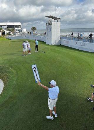Golf tournament at the seaside: players on a manicured green under cloudy skies, officials and signage near a viewing platform and ocean.