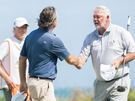On a sunny golf course, three men in golf attire exchange a handshake during a match.