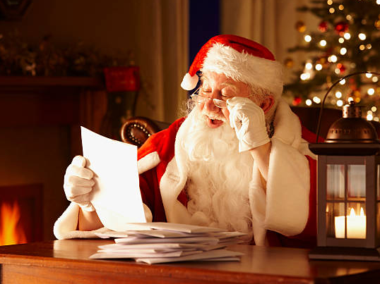 Santa Claus in a red hat reading a letter by a roaring fireplace, with stacked mail and a lantern beside him.