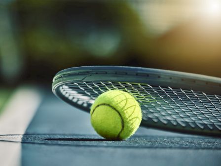 A tennis ball rests on a court near a racket, with green grass and a blurred background.
