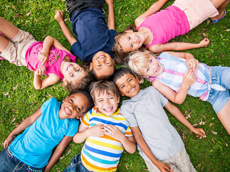 A group of kids lie together on green grass, smiling and looking up at the camera in a sunny outdoor moment.