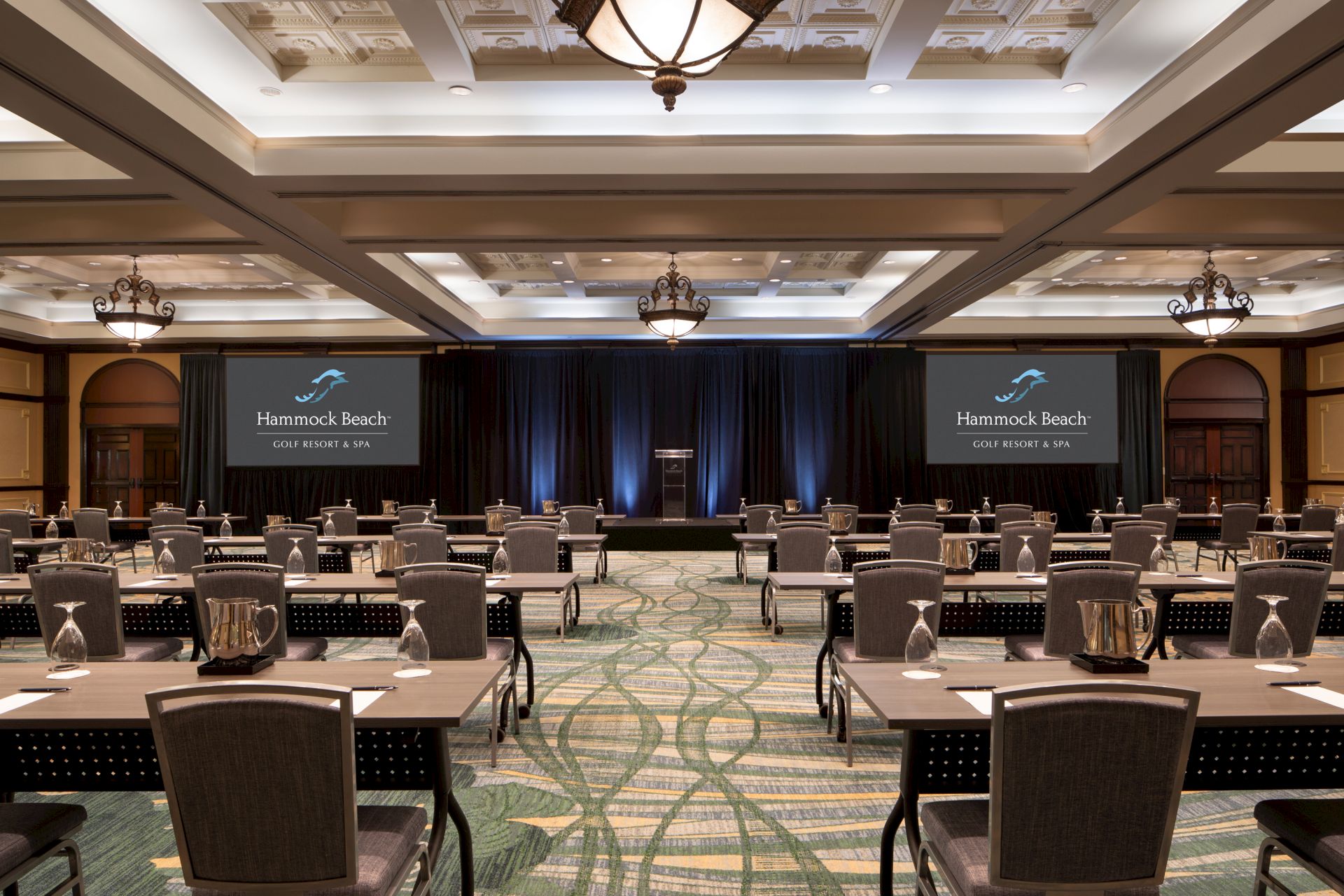 A conference room with rows of tables and chairs, two screens with "Homestead Beach" displayed, and ceiling lights.