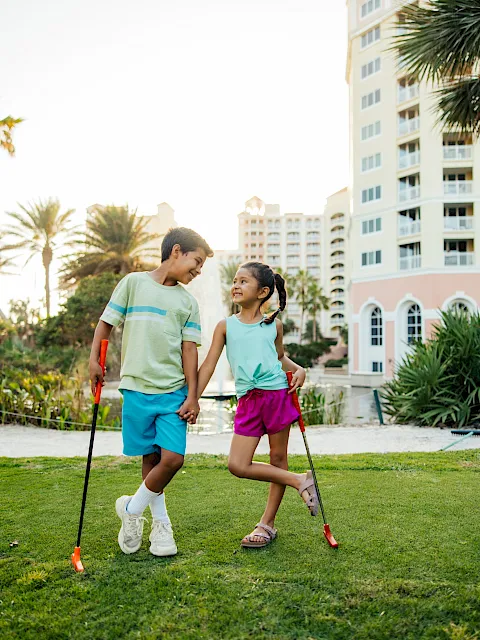 A boy and a girl play together on a sunny lawn with toy water guns, smiling by a hotel building.