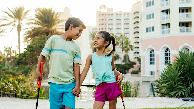 A boy and a girl play together on a sunny lawn with toy water guns, smiling by a hotel building.