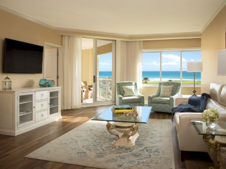 A bright seaside living room with two sofas, a coffee table, a TV cabinet, and large doors opening to an ocean view.