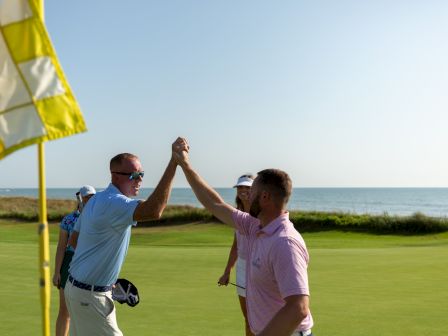 A golfer celebrates with a fist bump on a sunny seaside course, with a yellow flag and ocean in the background.
