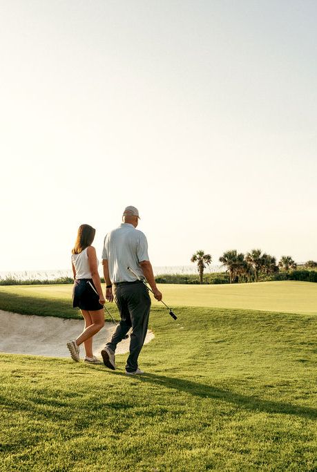 A man and woman walk hand-in-hand on a sunny golf course fairway with golf clubs.