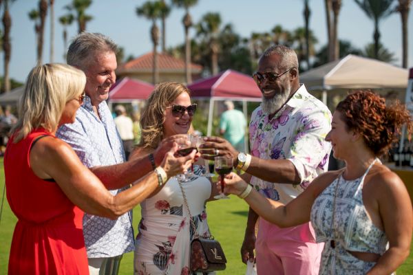 A group of five people outdoors clinking glasses, smiling, and wearing summer clothes, with tents and palm trees in the background.