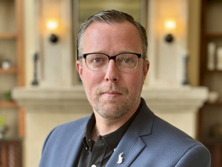 A man in a navy blazer and black shirt, wearing glasses, stands indoors with a neutral expression, softly lit background.