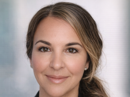 A professional headshot of a woman with light brown hair in a low ponytail, wearing a black blazer and white top, neutral expression.