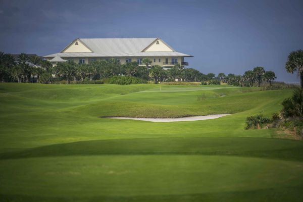 A manicured golf course with a large building in the background, surrounded by palm trees under a clear blue sky.