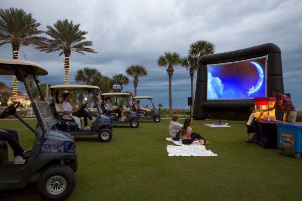 An outdoor movie screening on a lawn with golf carts, picnic blankets, and an inflatable screen, surrounded by palm trees under an overcast sky.