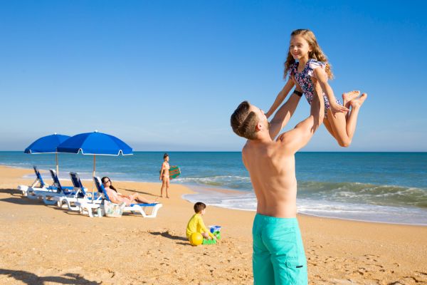 A beach scene with a man lifting a smiling child, people relaxing under blue umbrellas, and a child playing in the sand near the water.
