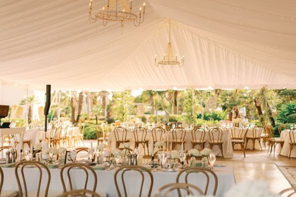 A beautifully decorated outdoor wedding reception with elegant chandeliers, white tablecloths, flowers, and chairs under a large tent.