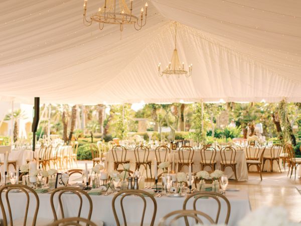 A beautifully decorated outdoor wedding reception with elegant chandeliers, white tablecloths, flowers, and chairs under a large tent.