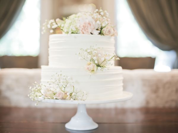 A two-tiered white cake adorned with delicate white and pink flowers on a white cake stand, in a softly blurred elegant setting.