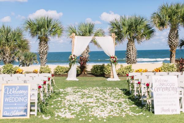 A beach wedding setup with white chairs, floral decorations, and a draped arch. Signs welcome guests to an unplugged ceremony.