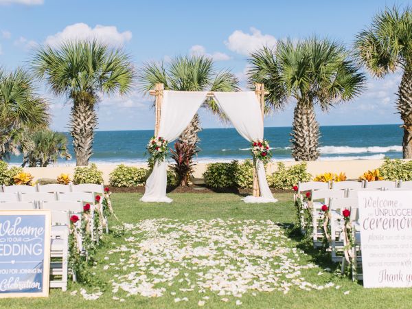 A beach wedding setup with white chairs, floral decorations, and a draped arch. Signs welcome guests to an unplugged ceremony.