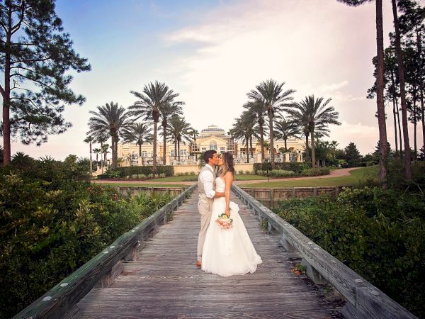 A couple in wedding attire kisses on a wooden bridge with a grand building and palm trees in the background.