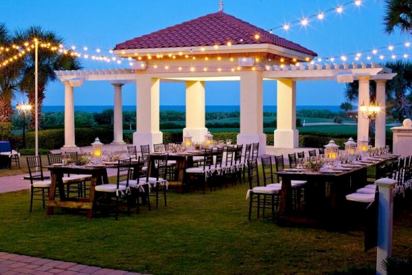 Outdoor dining setup with string lights and a gazebo, set on a lawn with tables and chairs, overlooking a scenic view.
