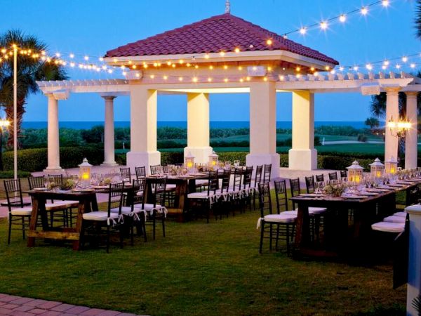 Outdoor dining setup with string lights and a gazebo, set on a lawn with tables and chairs, overlooking a scenic view.