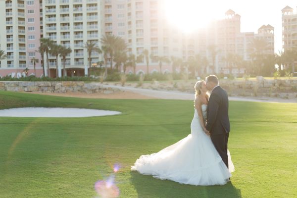 A couple in wedding attire stands on a golf course in front of tall buildings, illuminated by warm sunlight.