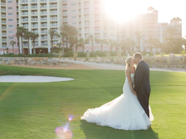 A couple in wedding attire stands on a golf course in front of tall buildings, illuminated by warm sunlight.