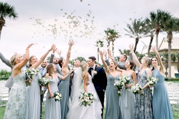 A joyful wedding scene with a bride and groom kissing, surrounded by a cheering bridal party tossing flower petals in the air.