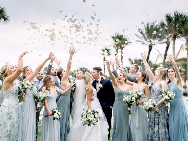 A joyful wedding scene with a bride and groom kissing, surrounded by a cheering bridal party tossing flower petals in the air.