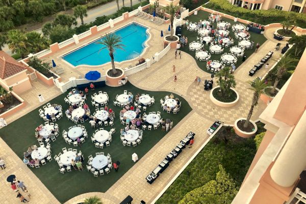 An outdoor event with round tables set on grass and a patio, next to a pool surrounded by palm trees and buildings.