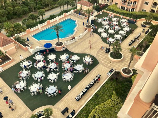 An outdoor event with round tables set on grass and a patio, next to a pool surrounded by palm trees and buildings.