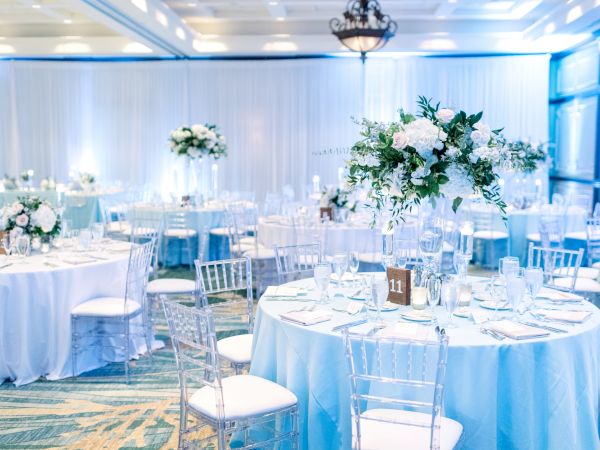 Elegant event setup with round tables, white tablecloths, floral centerpieces, and clear chairs in a well-lit room.
