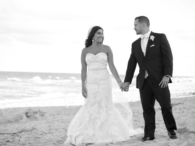 A bride and groom hold hands on a beach, smiling at each other, with the ocean in the background.