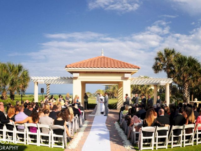 A beachside outdoor wedding ceremony with guests seated around an aisle. The bride and groom stand under a gazebo, surrounded by palm trees.