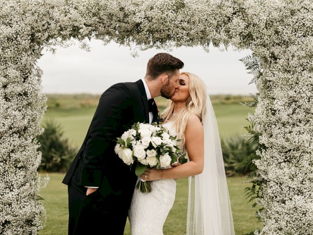A couple is kissing in front of a floral arch. The bride holds a bouquet of white roses, and they are outdoors.