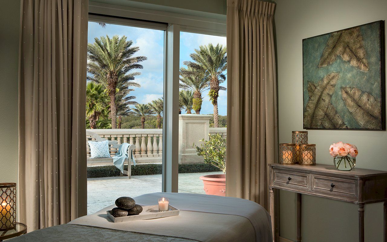 A tranquil room with a massage table, candles, and artwork, opening to a patio with palm trees through sliding glass doors.