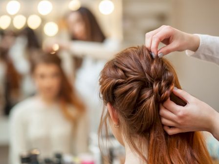 A hairstylist is braiding a woman's hair in a salon, with a mirror reflecting their image, creating a cozy and professional atmosphere.