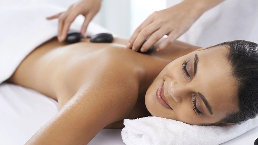 A person receives a relaxing hot stone massage while lying on a massage table, covered partially by a towel, with a smile of contentment.