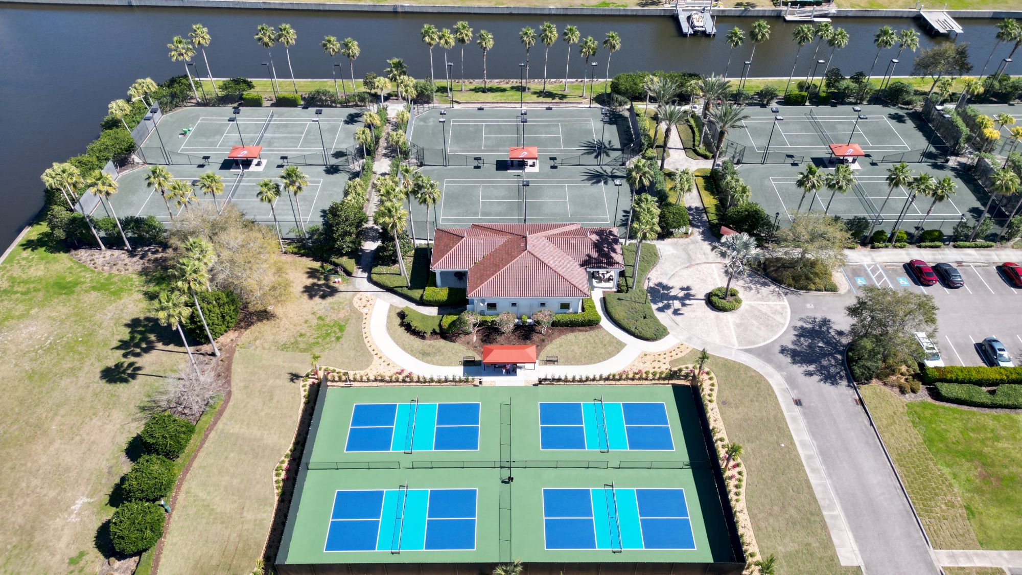 The image shows a sports facility with multiple tennis courts, a central building, parking area, and a nearby waterway, surrounded by trees.